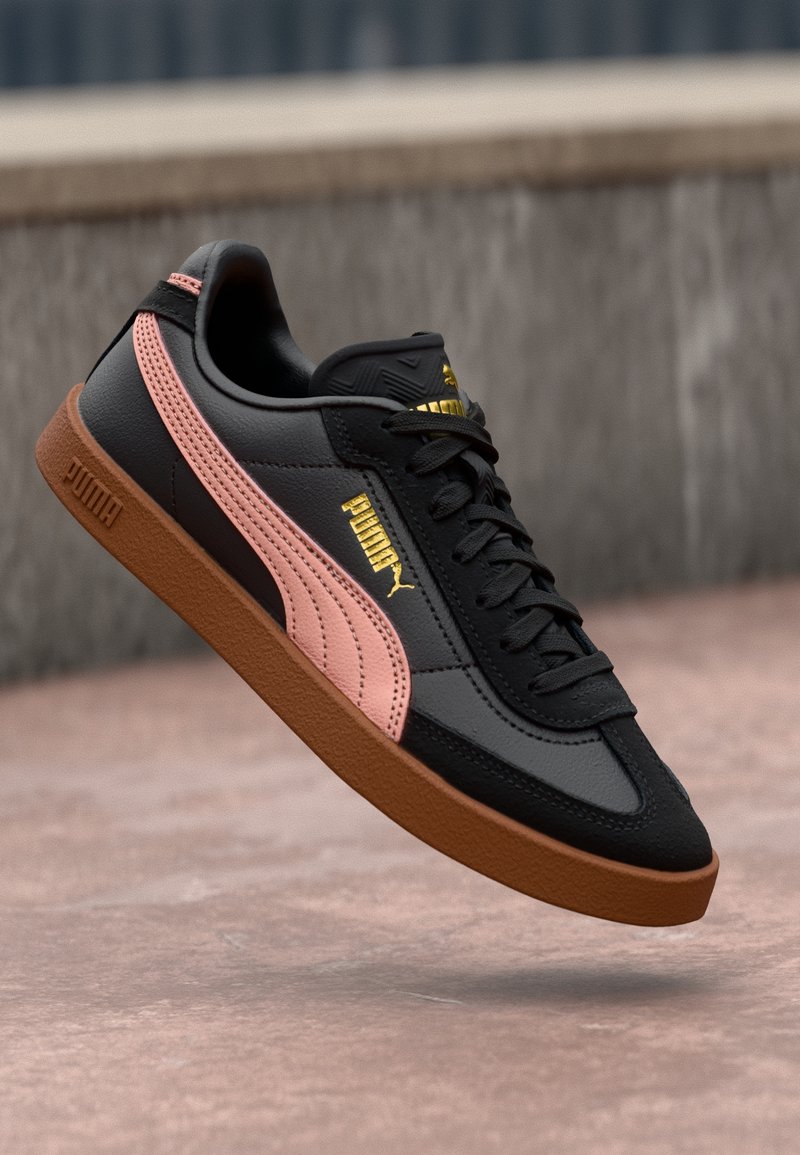 Baskets Puma noires avec une bande rose et une semelle marron flottant au-dessus d'une surface pavée avec un mur en béton flou en arrière-plan.