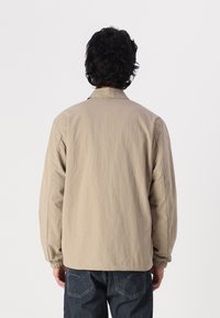 Chaqueta de color beige claro con cuello, puños elásticos y un corte relajado. Llevada sobre vaqueros oscuros, mostrando una textura suave y mate.