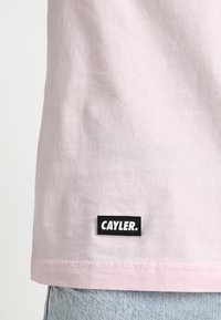 Camiseta de algodón de color rosa claro con una etiqueta rectangular negra que lleva el texto "CAYLER." situada cerca del dobladillo. Textura suave, diseño casual.