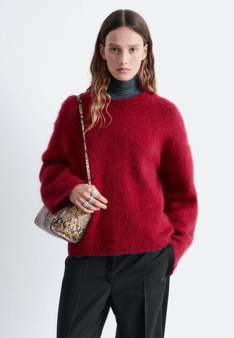 Pull rouge duveteux avec des manches surdimensionnées, associé à un sac à main à motif peau de serpent. Pantalon noir taille haute et col roulé noir en dessous.