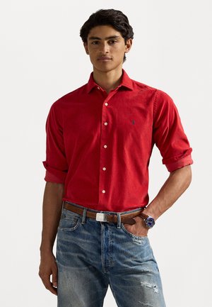 Rotes Hemd mit Knöpfen aus weichem Material, verziert mit einem kleinen blauen Logo auf der Brust, kombiniert mit abgenutzten Jeans aus Denim und einem braunen Gürtel.