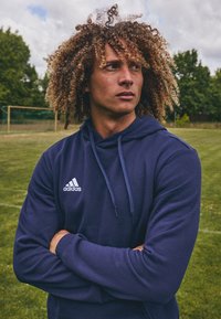 Marineblå hettegenser laget av mykt materiale, med en frontlomme i kengurustil og hvit Adidas-logo. Modellen står med armene krysset på en gresslette.