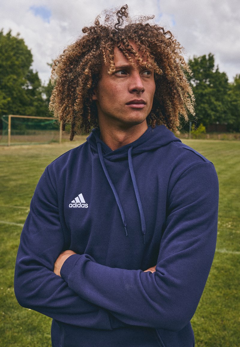 Marineblå hettegenser laget av mykt materiale, med en frontlomme i kengurustil og hvit Adidas-logo. Modellen står med armene krysset på en gresslette.