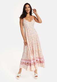 Maxi dress in light pink fabric featuring floral patterns, a V-neckline, and a tiered skirt with a decorative hem. Paired with white platform sandals.