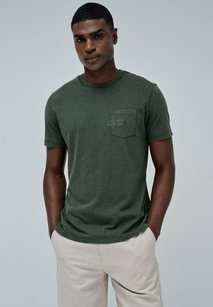 Camiseta de manga corta en verde oliva con escote redondo y un bolsillo en el pecho. La tela parece suave, con una textura sutil y un minimalismo en la marca.