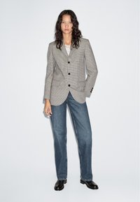 Vrouw met golvend haar die een geruite blazer, blauwe jeans, zwarte laarzen en een ketting draagt, staand tegen een effen witte achtergrond.