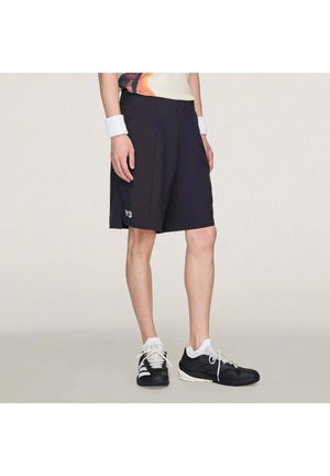 Person trägt schwarze Y3-Shorts, weiße Handgelenkbandagen und schwarz-weiße Sneaker, steht vor einem unifarbenen Hintergrund.