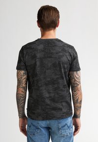 Černé tričko s krajinným vzorem, krátké rukávy, kulatý výstřih, vyrobené z měkké látky. Kombinováno s modrými džíny, ukazující tetovanou ruku.