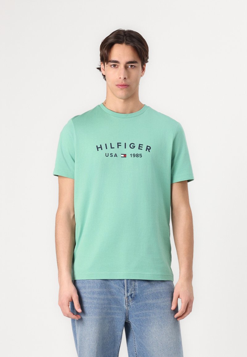Mintgrønn bomull T-skjorte med korte ermer og rund hals. Har marineblå teksten "HILFIGER USA 1985" med en liten logoaksent.