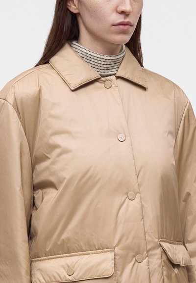 Femme portant une veste matelassée beige avec fermeture à boutons et poches à rabat, vue des épaules jusqu'au milieu du torse, sur fond blanc.