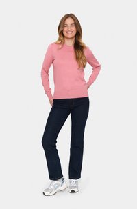 Ljusröd, ribbad långärmad tröja, kombinerad med mörka jeans och grå sneakers. Modellen står med händerna på höfterna och ler.