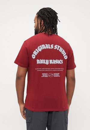 Mann mit Dreadlocks, der ein rotes T-Shirt trägt, auf dem in weißer Schrift "Originals Studio Daily Basics" steht, sowie zusätzlichem kleinerem Text auf der Rückseite.