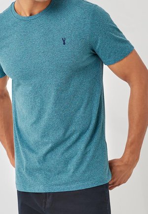 Mężczyzna w krótkim turkusowym t-shircie z małym ciemnoniebieskim logo jelenia na lewej stronie klatki piersiowej, w połączeniu z ciemnymi spodniami.