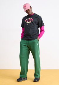 Zwarte Nike T-shirt met kleurrijk logo, roze lange mouw onderlaag, groene sweatpants en zwarte sneakers. Roze pet met "D" logo.