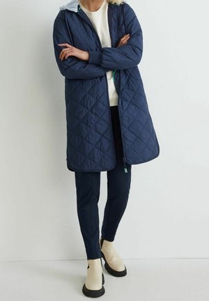 Persona vestida con un abrigo largo acolchado azul marino, suéter crema, pantalones azul marino y botines beige, de pie con los brazos cruzados.