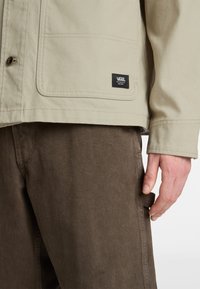 Beige canvas jacket with a front pocket and black Vans logo, worn over brown textured pants, with a hand gripping the pant's side.