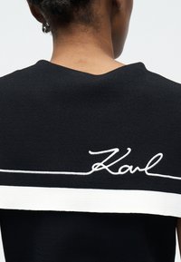 Πίσω όψη ενός ατόμου που φοράει μαύρο ρούχο με μια λευκή κεντητή γραμμή και το όνομα «Karl» στο πάνω μέρος της πλάτης.