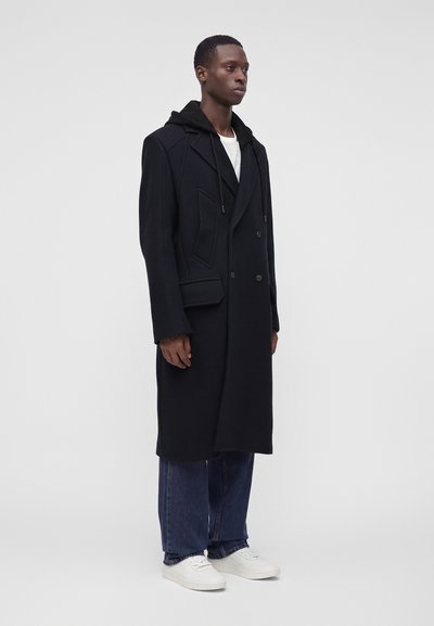 Homme portant un long manteau noir croisé sur un sweat à capuche noir, un jean bleu et des baskets blanches, debout devant un fond gris uni.