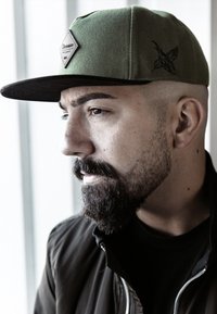Cappellino verde con visiera piatta, decorato con una toppa a forma di diamante contenente testo, dettagli neri e un design ricamato di lato. Indossato con una giacca nera.