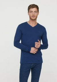 T-shirt à manches longues navy avec un col en V et une coupe ajustée. Texture douce, arborant un petit logo sur la poitrine. Assorti avec un jean bleu.