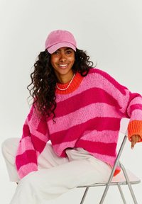 Živě pruhovaný svetřík v růžových a fuchsiových odstínech s texturovaným povrchem a oranžovým límcem. Doplněný o světlý růžový šátek a bílé kalhoty.