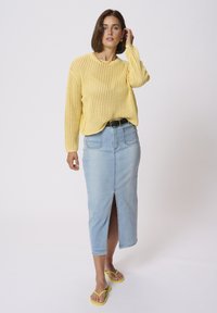 Suéter de punto amarillo con un corte holgado, combinado con una falda de mezclilla azul clara con una abertura frontal y chanclas amarillas.