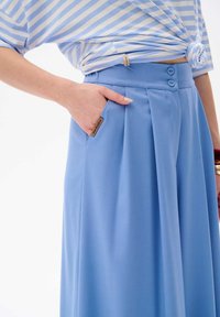 Lichtblauwe wijde broek met plooien, voorzien van een hoge taille, twee zijzakken en knoopsluiting. Gladde stof met een matte afwerking.