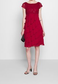 Czerwona sukienka z koronkową górą ozdobioną cekinami, z warstwowymi falbanami na spódnicy, krótkimi rękawami i długością do kolan. W komplecie z czarną kopertówką.