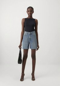 Γυναίκα με πλεξούδες, φορώντας μαύρο αμάνικο τοπ, τζιν σορτς ψηλής μέσης, ασημένιες γόβες και κρατώντας μια μαύρη τσάντα.