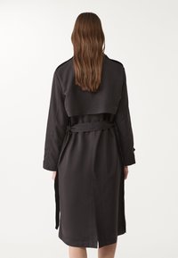 Schwarzer Trenchcoat mit taillierter Passform, langen Ärmeln und Gürtel in der Taille. Verfügt über einen verkürzten Vorderteil und seitliche Schlitze. Glatte Stofftextur.