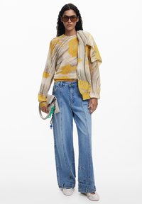 Gebreide trui in beige en geel, gecombineerd met hoge blauwe jeans en een grijze tas met kleurrijke bedels. Accessoires zijn onder andere een paar bruine zonnebrillen.