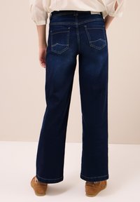 Persona vestida con jeans de pierna ancha azul oscuro y zapatos beige, vista desde la parte posterior contra un fondo beige claro.