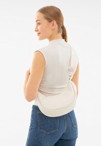 Bílá kožená crossbody taška s zakřiveným tvarem, zipovým uzávěrem a nastavitelným popruhem. Taška má jemný embosovaný detail loga.