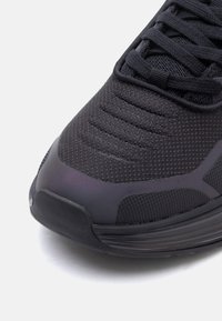 Krupan plan crne sportske obuće s prstom izrađenim od teksturiranog pletenog materijala, gumenim potplatom i čvrsto zavezanom vezicom na bijeloj pozadini.