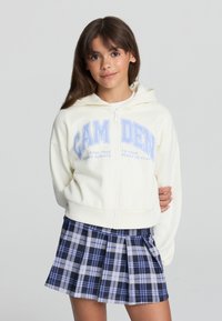 Felpa con zip di colore crema con scritte azzurre e una tasca frontale, abbinata a una gonna plissettata a quadri nei colori navy, azzurro e bianco.