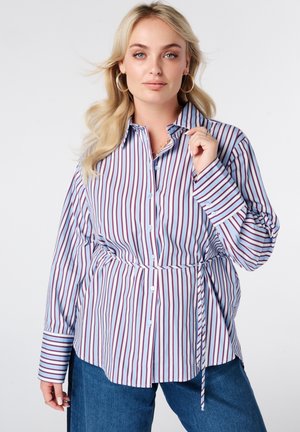 Femme aux longs cheveux blonds portant des boucles d’oreilles anneaux, une chemise boutonnée à rayures verticales bleues et rouges avec un lien à la taille, et un jean bleu.