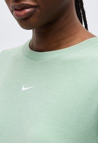 Κοντινό πλάνο ενός ατόμου που φοράει ένα ανοιχτό πράσινο μπλουζάκι με στρογγυλή λαιμόκοψη και μικρό λευκό λογότυπο Nike στη λογότυπο στο στήθος.