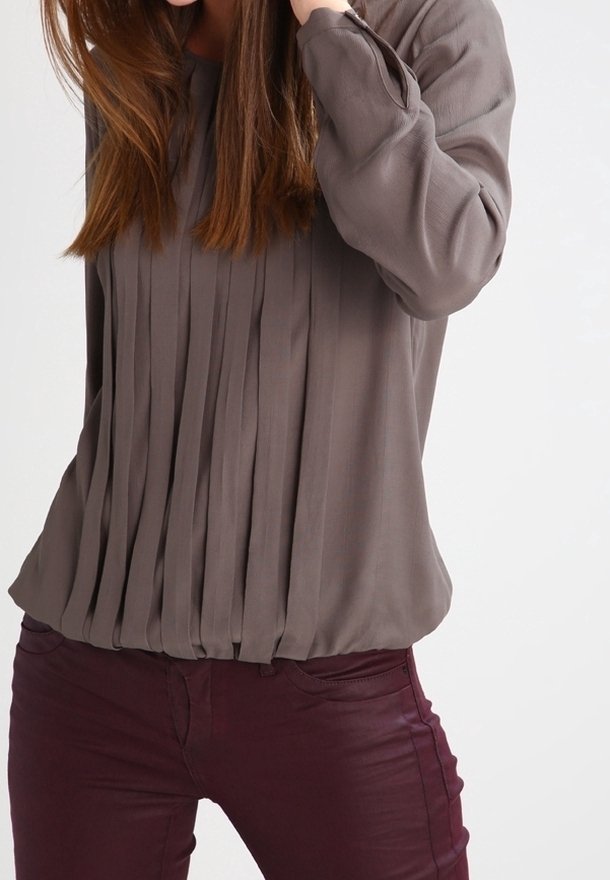 Blouse à manches longues plissée de couleur grise douce, confectionnée dans un tissu doux. Associée à un pantalon ajusté bordeaux, créant un contraste texturé.
