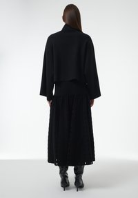 Vrouw met lang bruin haar draagt een zwarte cropped sweater, een gestructureerde zwarte midi-rok en zwarte haklaarzen, staande op een witte achtergrond.