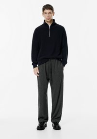 Fiatal férfi áll, fekete fél cipzáros pulóverben, laza sötétszürke nadrágban és fekete bőrcipőben egy fehér háttér előtt.