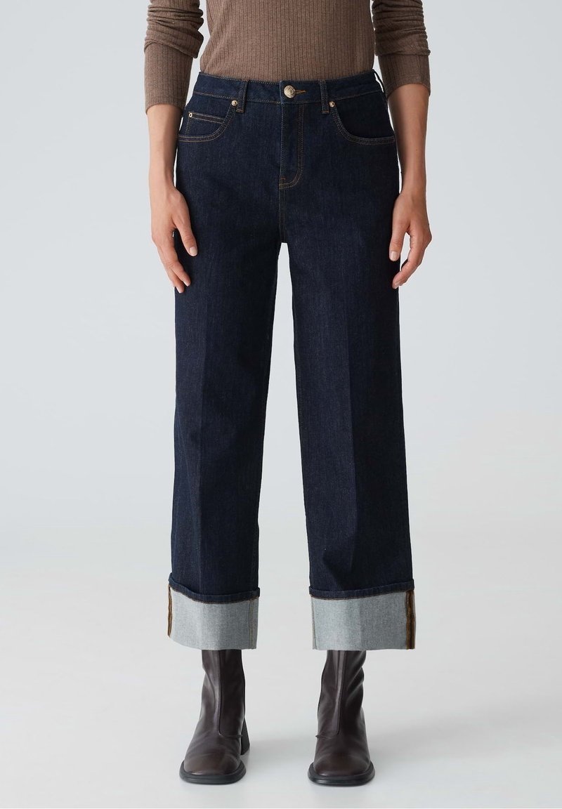 Femme portant un jean large à pattes d'éléphant bleu marine avec des revers, un haut en ribbed marron à manches longues et des bottines à talon marron foncé, se tenant devant un fond uni.