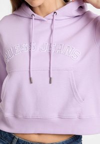 Sudadera cropped en lavanda con bolsillo frontal, logo bordado "GUESS JEANS", cordones con puntas metálicas y tejido texturizado.