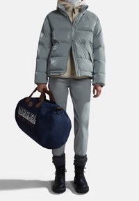 Grå pufferjakke med lynlås foran, beige hoodie nedenunder, grå bukser, sorte snørresko og en navy duffeltaske med logoaccentuer.