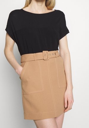 Woman wearing a black short-sleeve top tucked into a high-waisted tan skirt with stitched pockets and a matching belt.