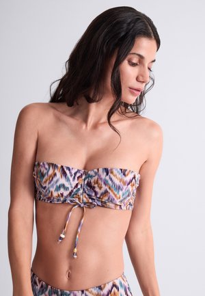 Femme aux longs cheveux foncés portant un haut de bikini bandeau sans bretelles à motif avec un cordon noué à l'avant, regardant vers le bas à sa gauche.