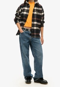 Camisa de franela a cuadros en negro, blanco y burdeos sobre un top corto naranja, combinada con vaqueros holgados azules y zapatos negros.