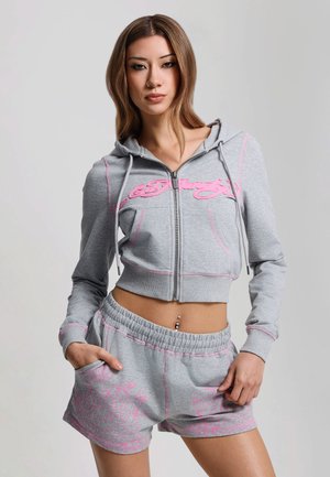 Jeune femme portant un sweat à capuche gris court zippé et un short assorti avec des motifs floraux roses, debout les mains dans les poches.
