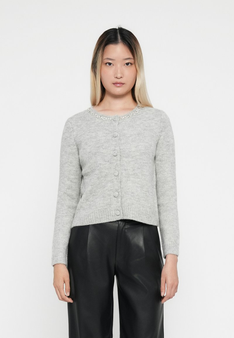 Cardigan en tricot gris avec un col rond, orné de passepoils et doté d'une fermeture à cinq boutons, porté avec un pantalon en cuir noir.
