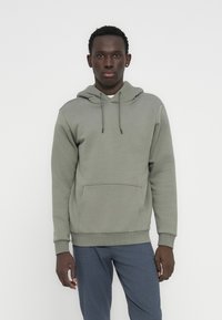 Νεαρός άνδρας στέκεται μπροστά σε λευκό φόντο, φοράει ανοιχτοπράσινο φούτερ με κουκούλα και μπλε παντελόνι, κοιτάζοντας μπροστά με ουδέτερη έκφραση.