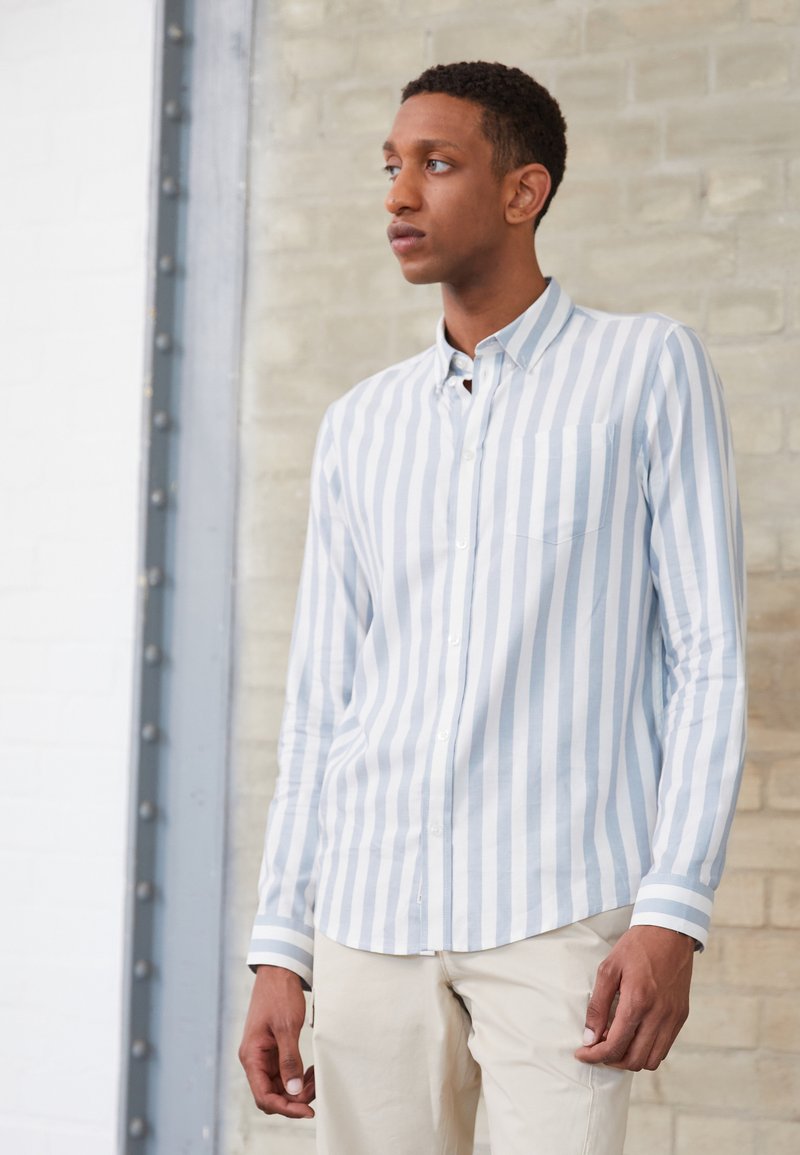 Camicia a righe blu chiaro e bianca con taschino sul petto, maniche lunghe e polsini a righe a contrasto. Indossata con pantaloni color crema.
